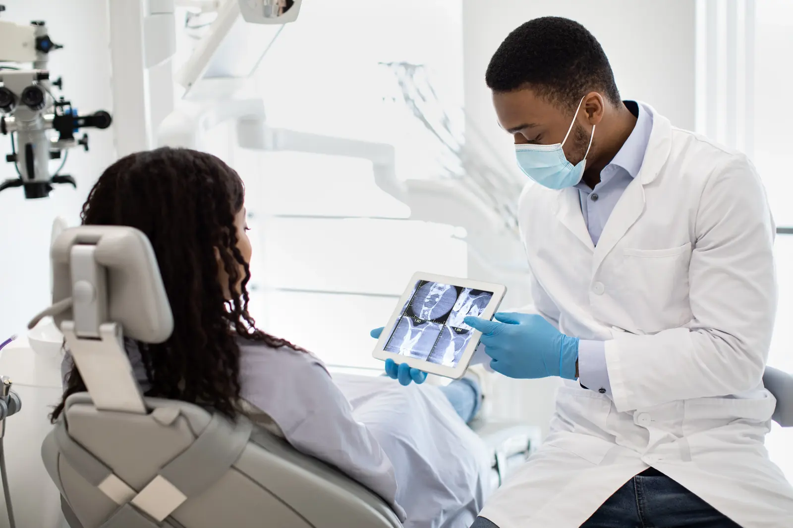 A masked dentist showing a patient in a dental chair an X-ray scan on a tablet