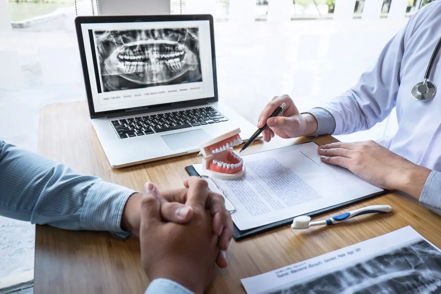 A dentist pointing a pen at a tooth model beside a laptop showing a dental X-ray, across the desk from a patient