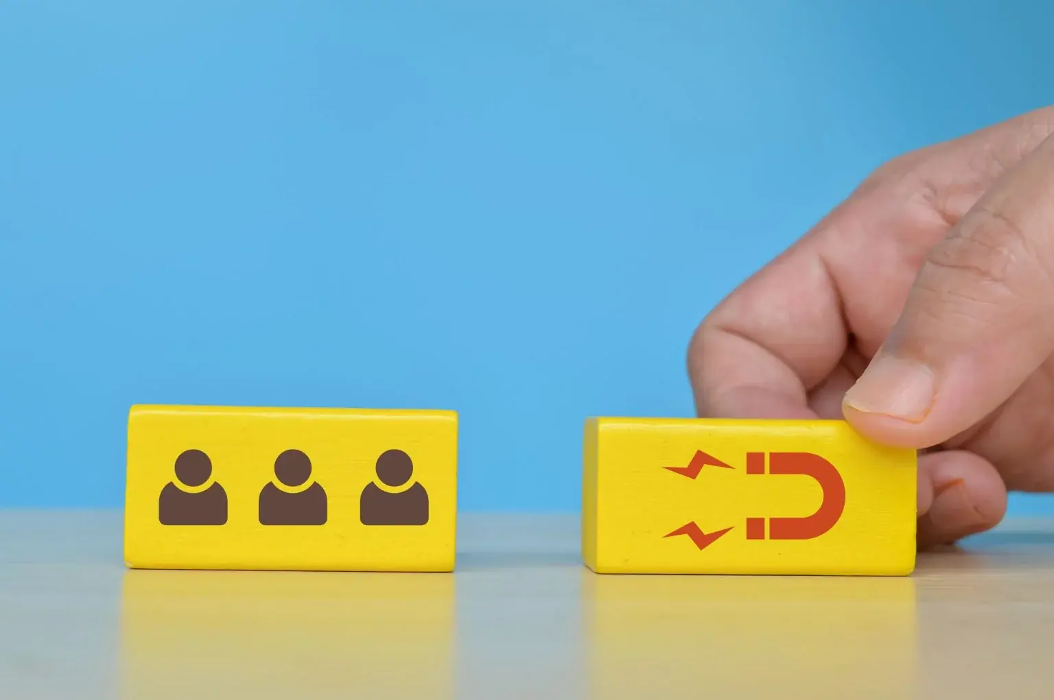 A hand placing a yellow wooden block printed with a magnet icon next to a block printed with three person icons, on a blue background