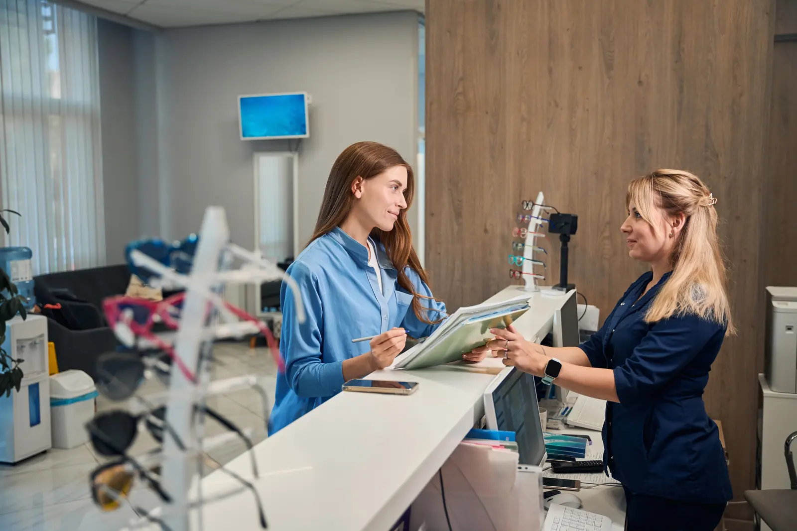 A patient holding a pen and paperwork talks with a staff member across a front desk, with eyeglass frames on a display stand in the foreground