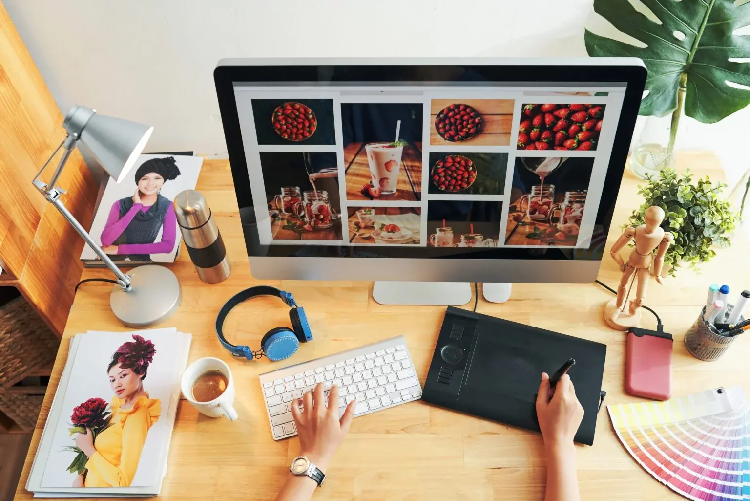 Overhead view of a designer’s desk: one hand on a keyboard and one on a pen tablet, in front of a monitor showing a grid of food photos