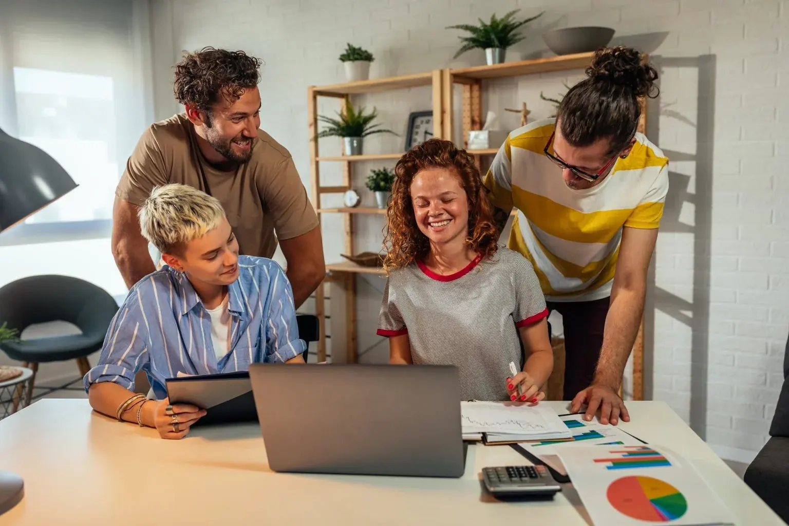Four smiling colleagues gathered around a laptop and printed charts at an office desk