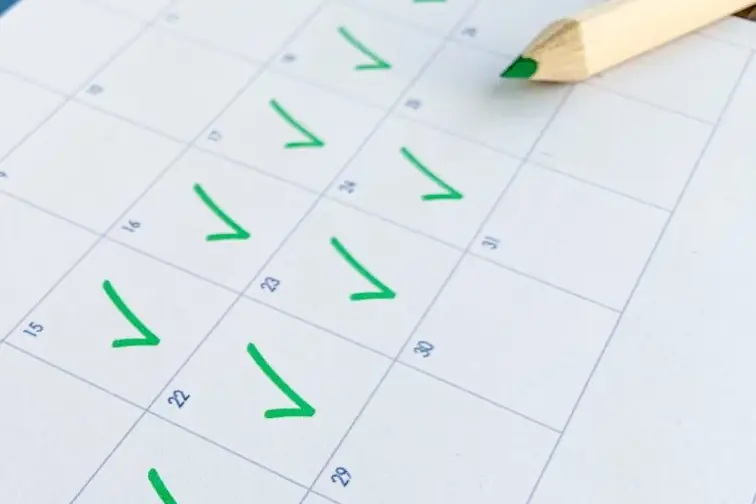 A printed calendar grid with green check marks on several days and a green-tipped pencil resting on the page